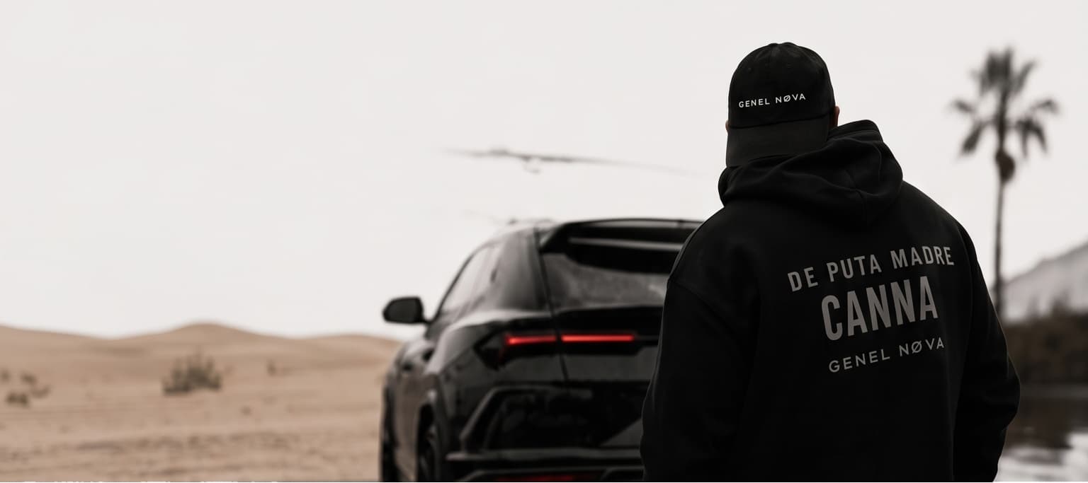 A figure in a De Puta Madre Canna hoodie facing a black car in the desert